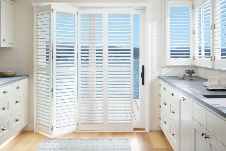 Transitional style kitchen with a glass sliding door revealing a patio and beach view, partially covered by Polysatin Shutters. Made in Australia. Water resistant. Available in Complete Blinds Brisbane.