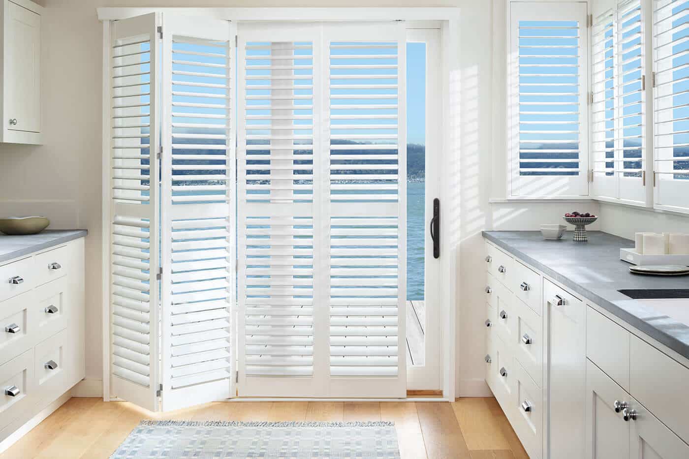 Transitional style kitchen with a glass sliding door revealing a patio and beach view, partially covered by Polysatin Shutters. Made in Australia. Water resistant. Available in Complete Blinds Brisbane.