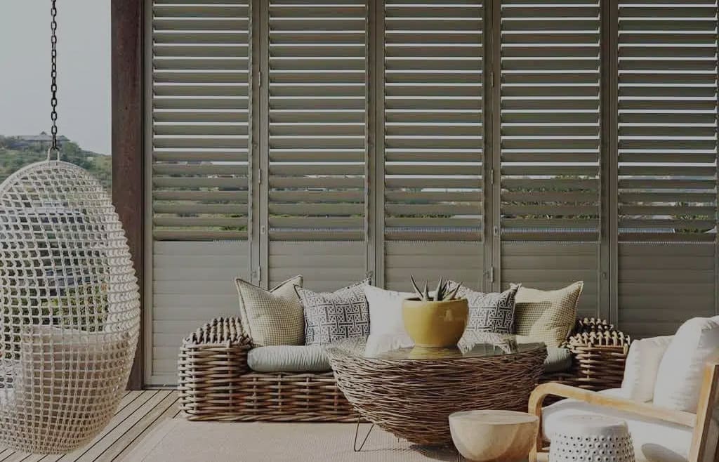 Light brown aluminium shutters in a eclectic-styled outdoor space/patio. Available in various panel arrangements. For sale in our Brisbane showroom.