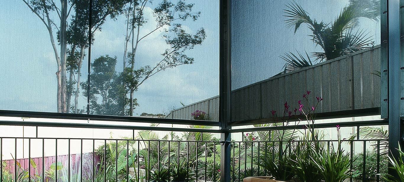current_media_luxaflex-products-external-collection-straight-drop-awnings-banner Home patio by the garden with flora and fauna, featuring sheer black straight drop awnings, allowing us to partially see the outside view. By Complete Blinds Brisbane.
