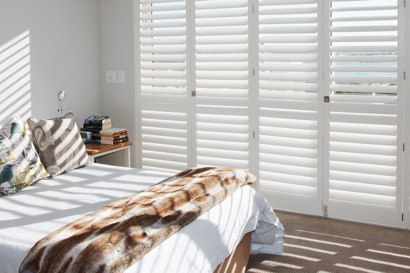 Cozy white bedroom with wooden floors installed with Brightwood Shutters, sunlight filtering through it, creating streaking light and shadow contrast. Available at Complete Blinds Brisbane.