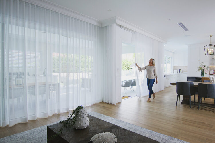 Woman adjusting sheer white curtains of her living room window, installed by Complete Blinds Brisbane.