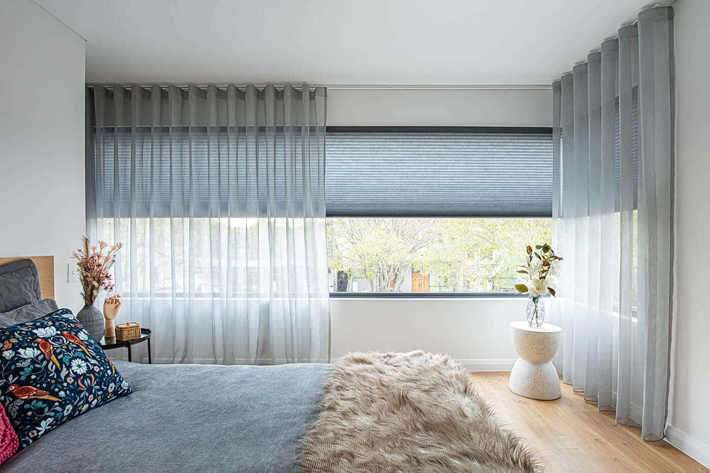 Contemporary aesthetic bedroom featuring windows installed with Duette shades and sheer grey curtain, for privacy and light control. Curtains for sale at Complete Blinds Cleveland.