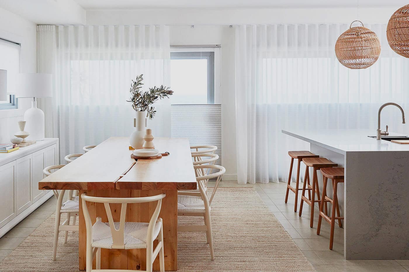 Aesthetic earth-tone kitchen with concrete, featuring windows installed with sheer white curtains- allowing light and privacy control. Available at Woman walking in her living room, while adjusting/closing her sheer white window curtains, for privacy and light control. Available at Complete Blinds Indooroopilly.