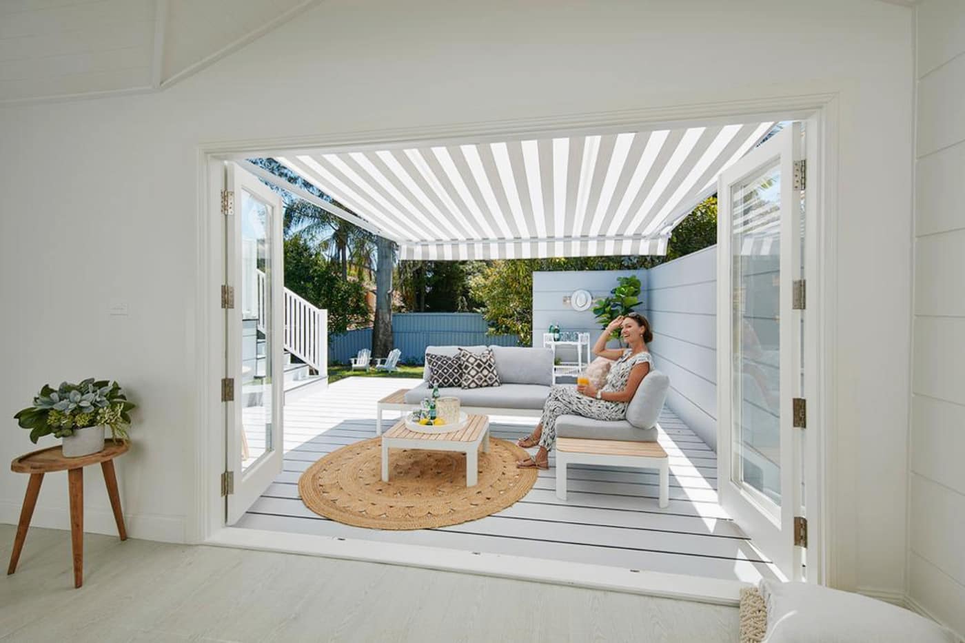 Luxury Blinds Epping Woman relaxing on couch in a white and grey bright Nordic home patio in Chermside installed with striped Folding Arm Awnings. View from inside.