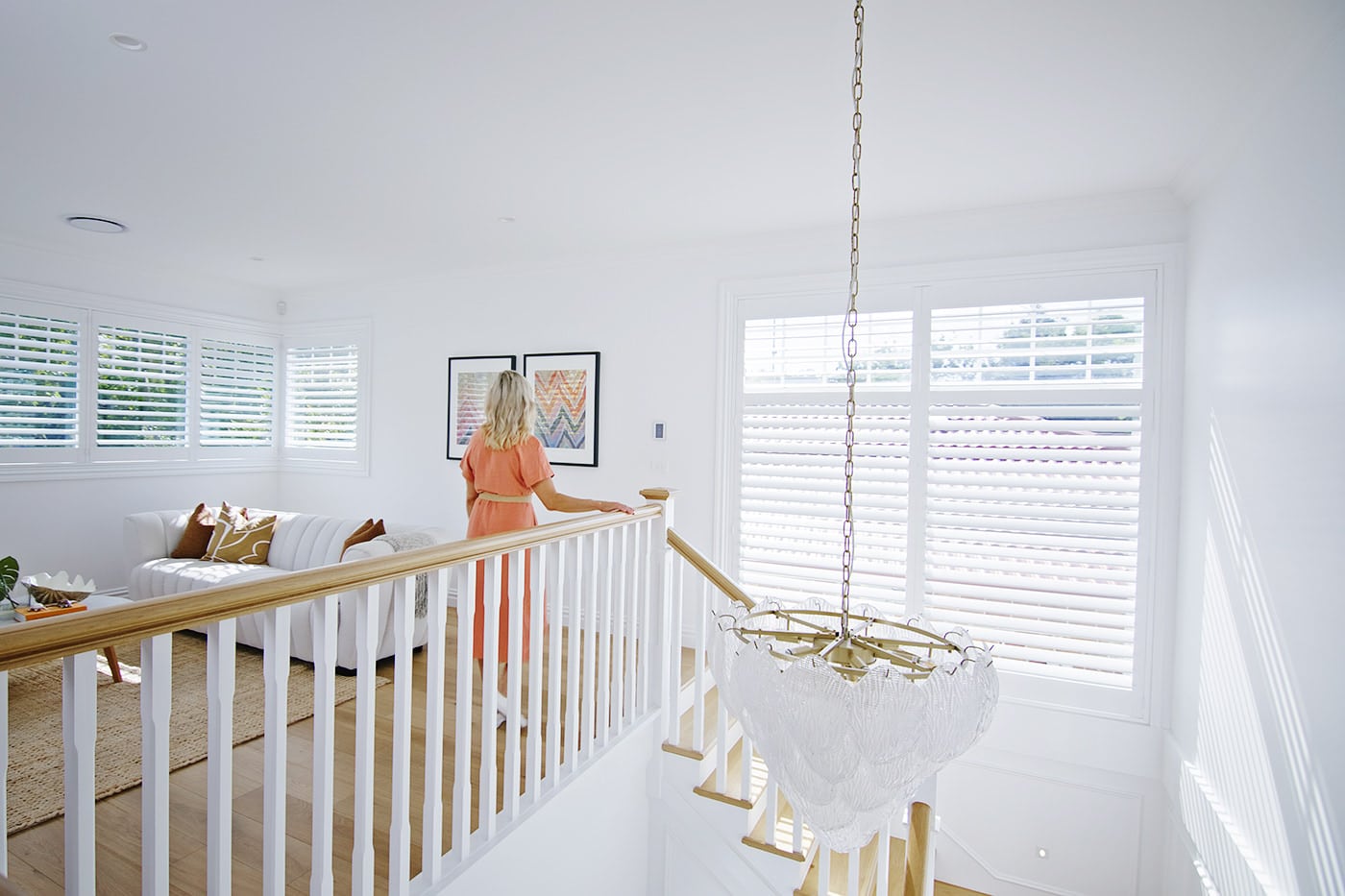 Luxury Blinds Epping Woman in orange dress by the stairs of a second-storey living area, featuring windows installed with white plantation shutters. Available at Complete Blinds Indooroopilly.