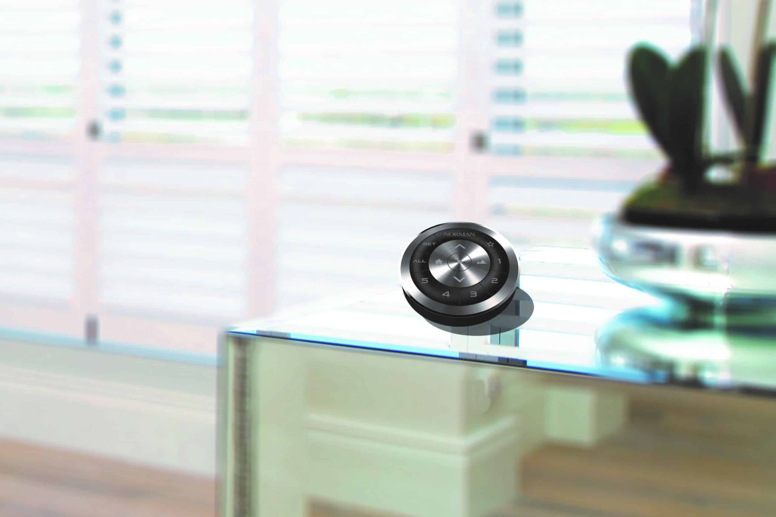 IMAGE 19 A sleek black remote Norman curtains and blinds control device sits on a modern glass table with a blurred background of white plantation shutters and wooden flooring. A plant in a glass bowl is partially visible on the right side of the image.