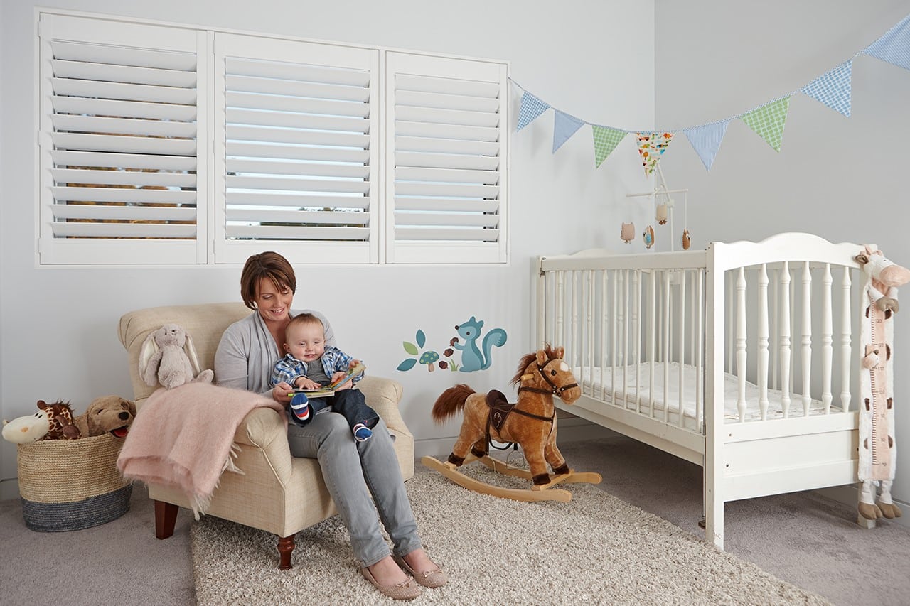 IMAGE15 Cozy nursery room with a mother sitting in an armchair reading a book to her baby. The room features white plantation shutters, a white crib with soft toys, a plush rocking horse, and a decorative wall mural of a squirrel. A string of colorful bunting hangs across the wall, and a soft pink blanket drapes over the armchair beside a basket of stuffed animals.