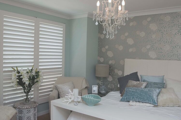 Elegant bedroom with soft pastel tones, featuring a floral accent wall, a chandelier with crystal details, and a white bed adorned with assorted textured pillows. The room includes a cozy armchair with a cushion and throw, white plantation shutters, and decorative accents like a vase of white lilies and a seashell-shaped bowl on a bedside table.